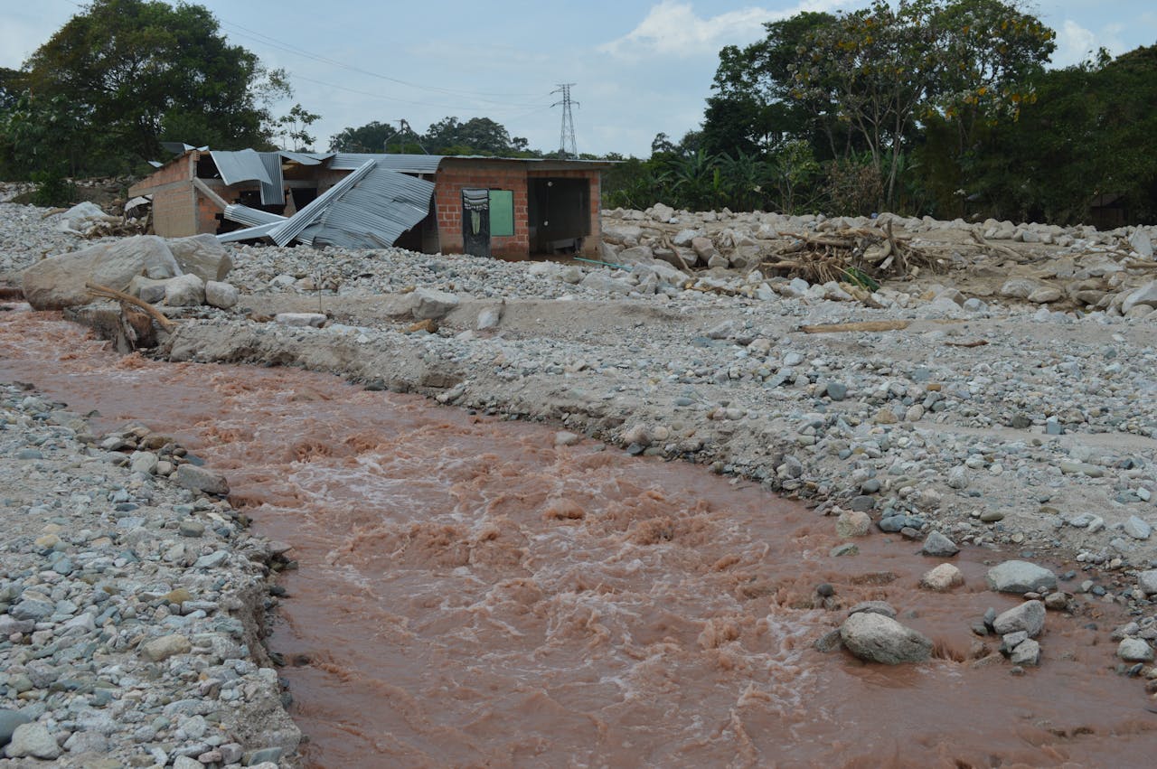 our-services-3 A damaged house stands near a muddy river after a flood, illustrating the impact of natural disasters.