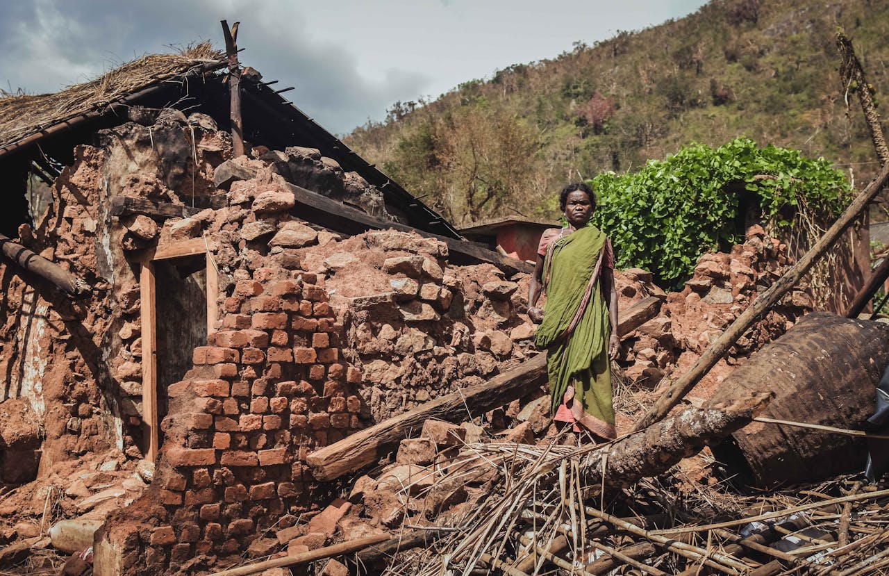 gallery-6 Woman stands in front of a destroyed house after a disaster in rural India, depicting struggle.